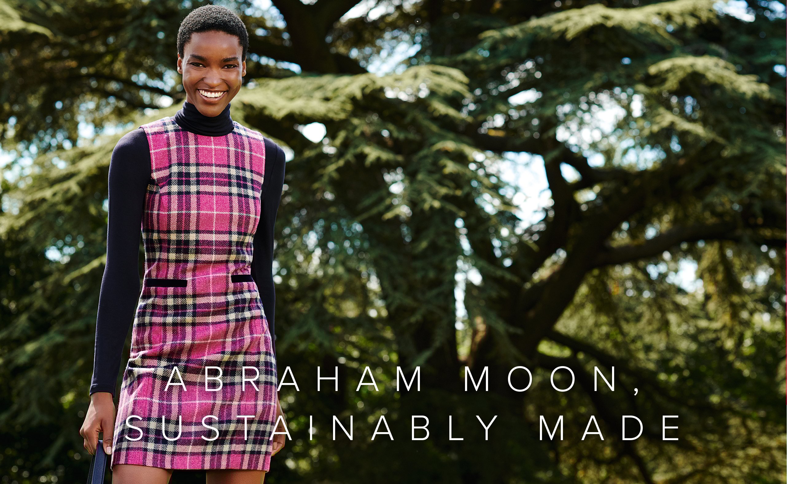 Alt text: Hobbs wool shift dress with a check pattern in pink shades made from Abraham Moon’s British made sustainable wool fabric, layered over a navy blue turtleneck women’s jumper styled with a leather tote bag.