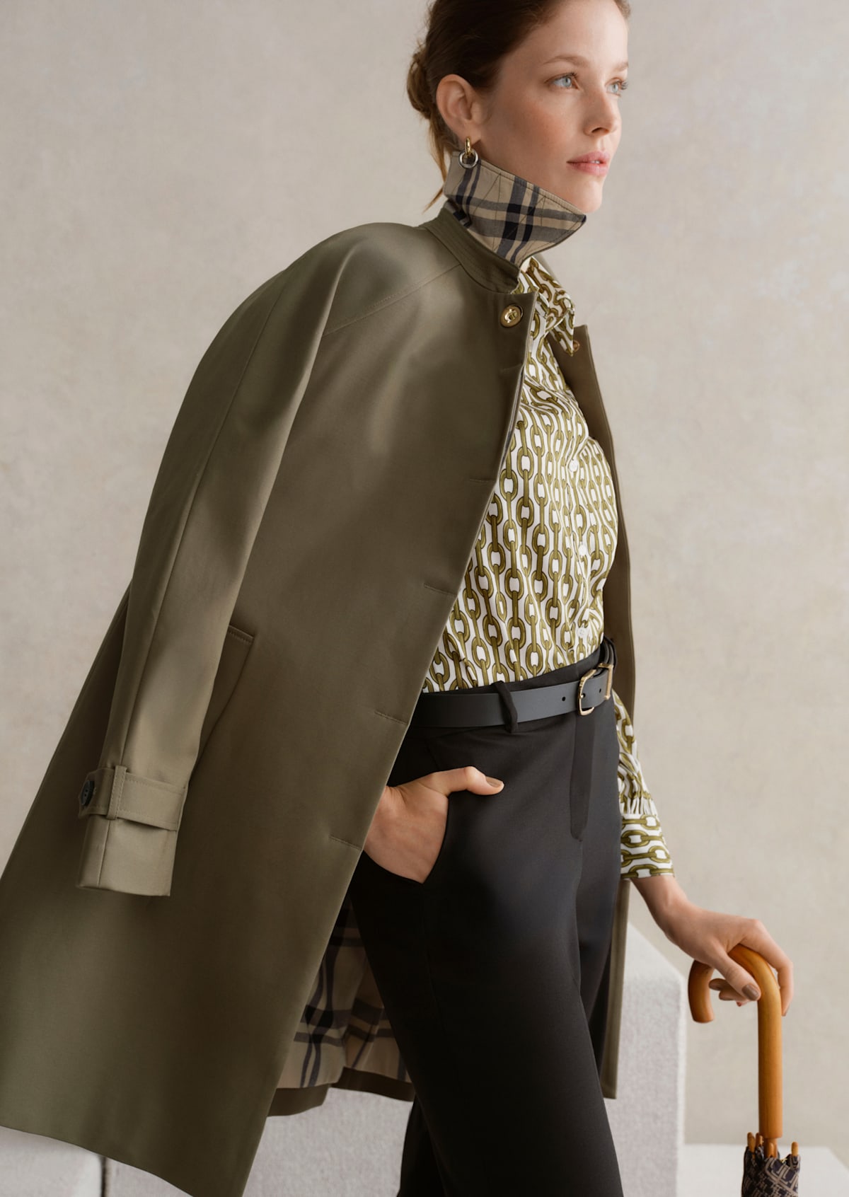 A gallery of two images displaying a model wearing a khaki trench coat, printed blouse and tailored trousers.