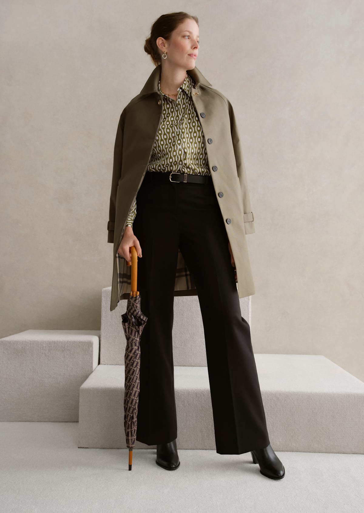 A gallery of two images displaying a model wearing a khaki trench coat, printed blouse and tailored trousers.