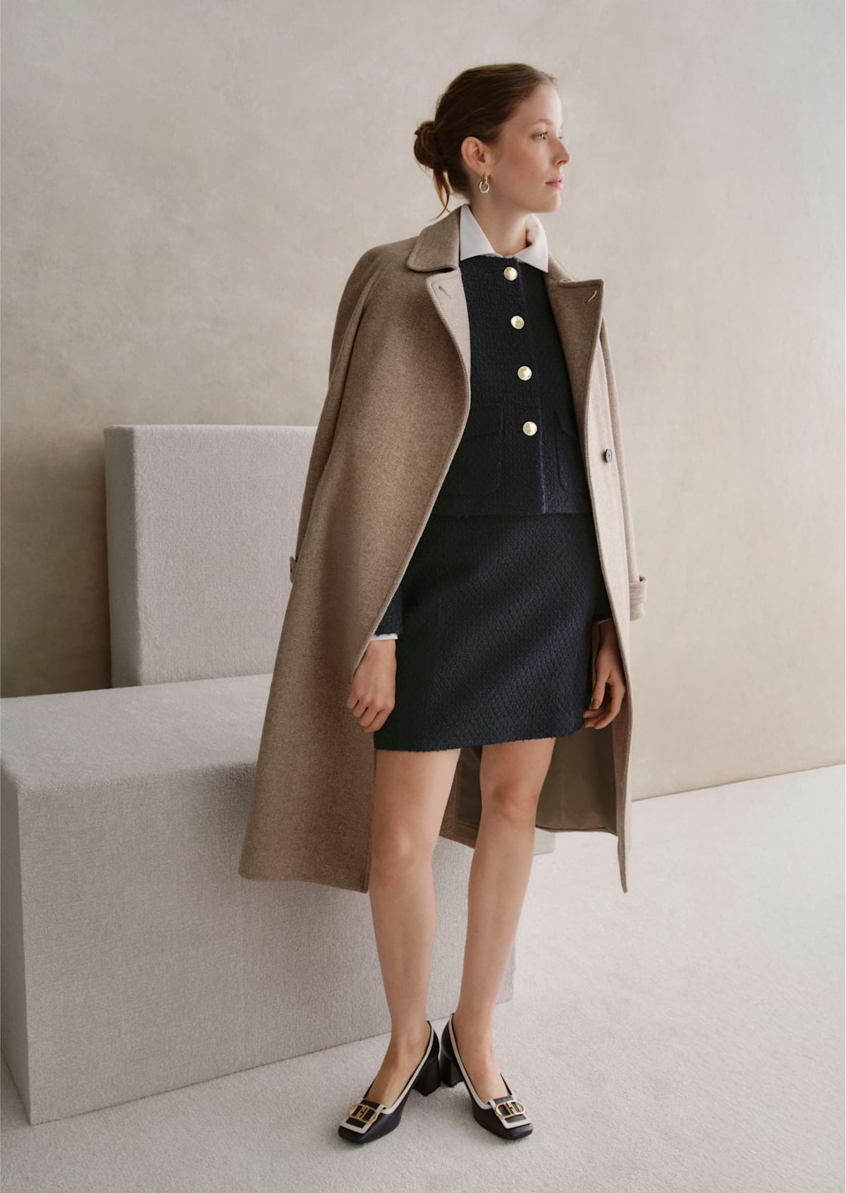 An image of a model wearing a beige tailored coat over a navy skirt and jacket