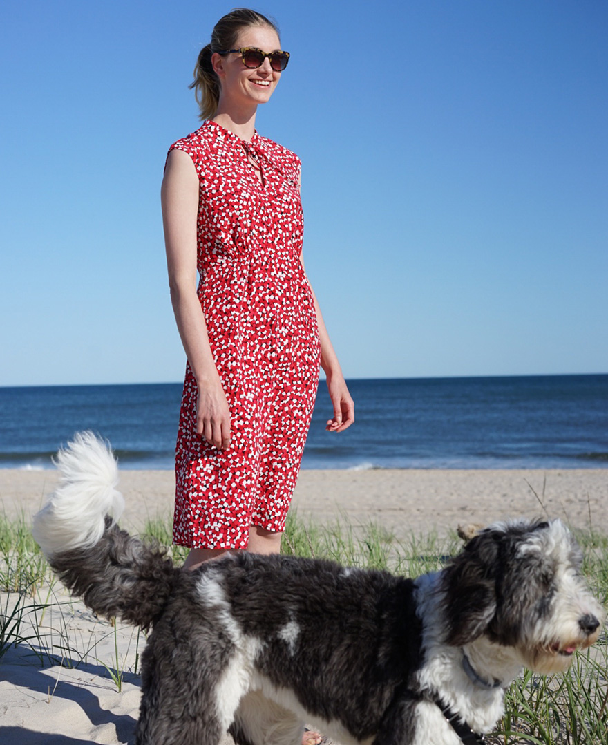 Hobbs model, Irish Van Berne,photographed on the beach in a red floral sleeveless dress with a grey and white dog. 