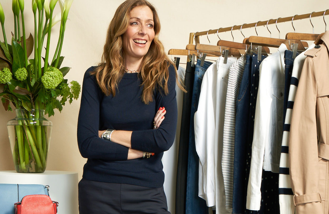 Kat Farmer photographed by a clothing rail of women's occasion dresses.