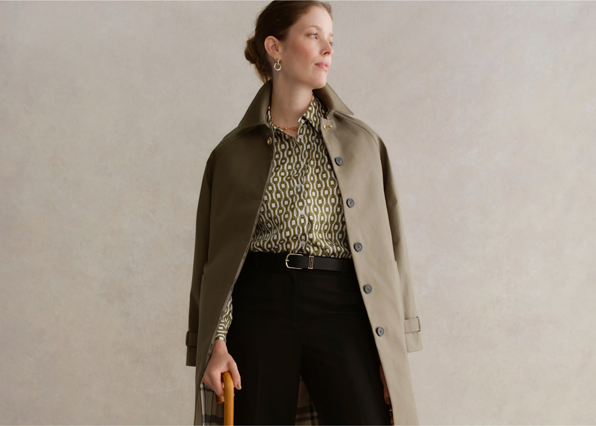 An image of a model wearing a khaki trench coat, printed blouse and tailored trousers.